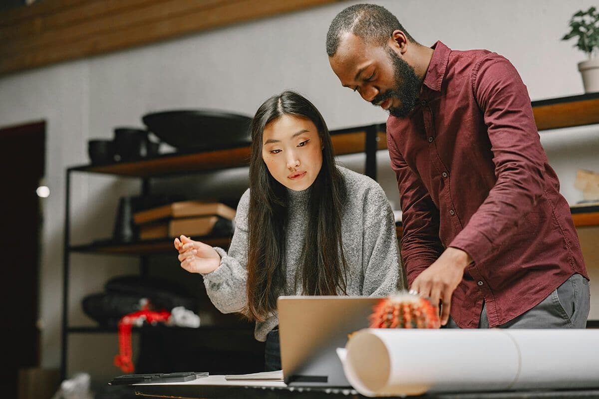 man and woman standing at a desk in an office looking at a laptop as they discuss collaborative culture at their company