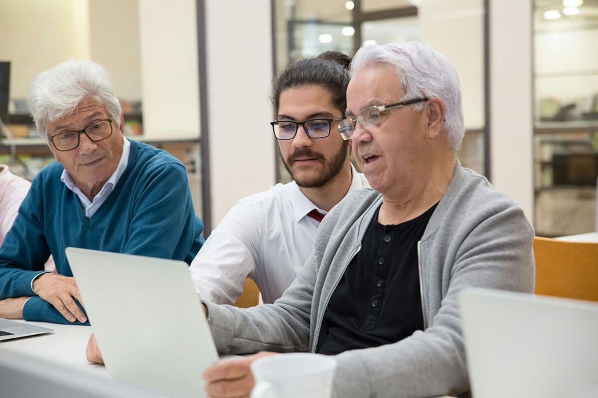 Three older men look at a laptop during a team mentoring meeting, learning about the company's new intranet system.